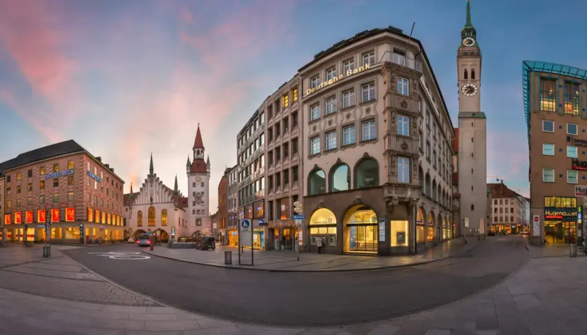 Abenddämmerung in Lehel, München. Die Deutsche Bank und das Ludwig Beck Gebäude erstrahlen im warmen Licht, während der Turm der Frauenkirche in den rosafarbenen Himmel ragt.