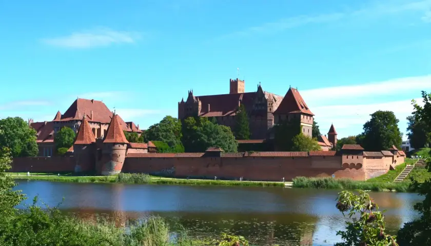 Potężny zamek Malbork z czerwonej cegły, otoczony murami obronnymi, góruje nad spokojną wodą.