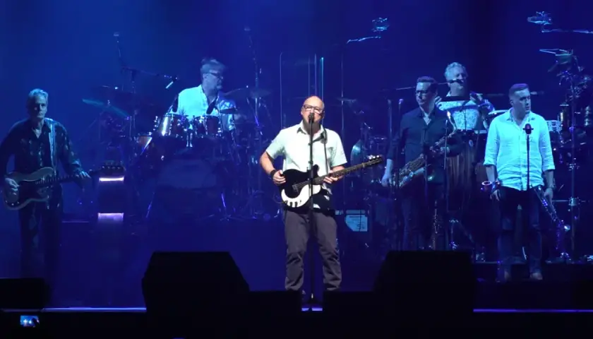 Mark Knopfler na scenie z zespołem podczas jednego z jego niezapomnianych koncertów. Muzycy grają na gitarach, saksofonach i perkusji.