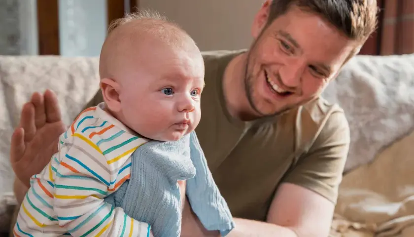 Padre sorridente tiene in braccio un neonato con una copertina azzurra. La posizione del neonato sembra confortevole, forse per alleviare il singhiozzo.