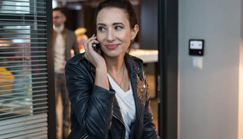 Aleksandra Popławska w skórzanej kurtce rozmawia przez telefon. W tle widać mężczyznę i biurowe wnętrze, przypominające scenę z jej filmów i seriali.