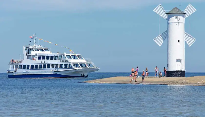Biała latarnia morska wiatrakowa, przypominająca młyn, stoi na piaszczystym cyplu. Obok cumuje biało-niebieski statek wycieczkowy. Kilka osób odpoczywa na brzegu. To jeden z urokliwych widoków na polskim wybrzeżu.