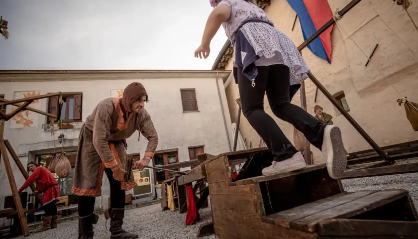 Bambini si divertono con giochi medievali. Un uomo in costume d'epoca aiuta una bambina a salire su una struttura di legno.