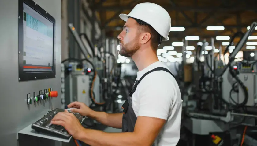 Operator maszyn CNC w białym kasku pracuje przy panelu sterowania, wprowadzając dane na klawiaturze.