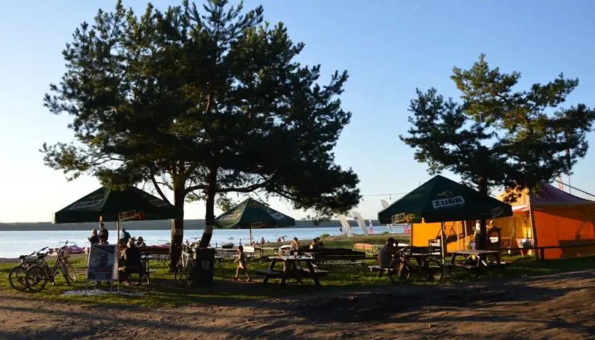 Relaks na plaży Karolinów. Ludzie siedzą pod parasolami, rowery czekają na plaży, a w tle jezioro i namioty.