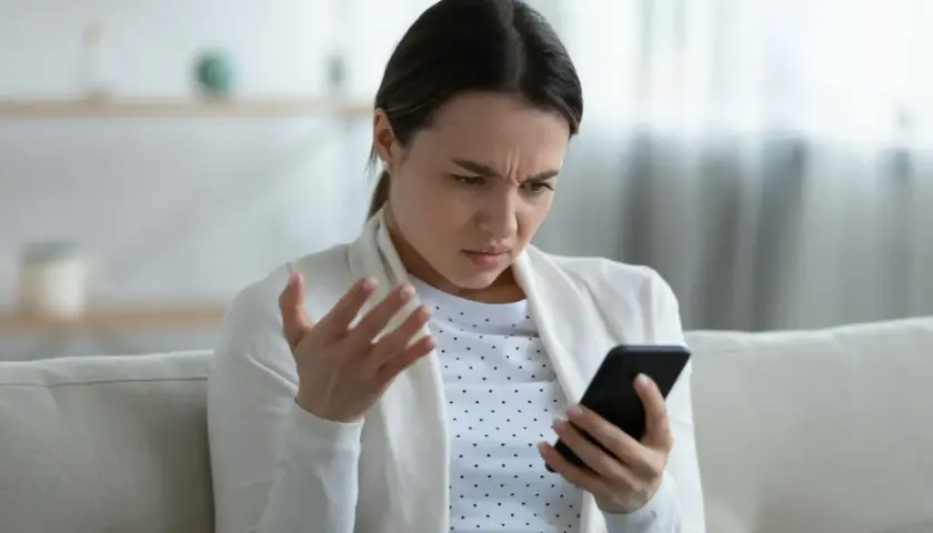 Kobieta z irytacją patrzy na telefon, gestykulując ręką. Ewidentnie przeszkadza jej słaby internet w telefonie, uniemożliwiający normalne korzystanie.