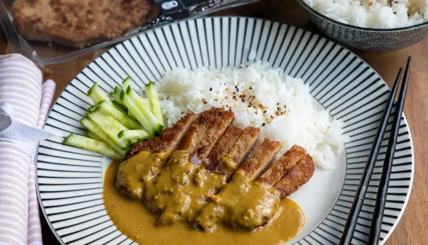 En tallrik med katsu curry, ris och gurka. Den panerade kotletten är skuren i skivor och täckt med en krämig currysås.
