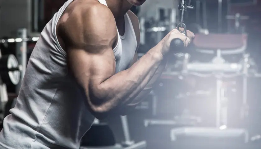 Mężczyzna ćwiczy mięsień trójgłowy na wyciągu. Widoczny jest jego umięśniony biceps i przedramię.