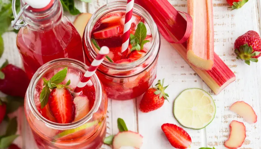 Orzeźwiający sok z rabarbaru z truskawkami i miętą, podany w słoikach z lodem i słomkami. Obok leżą świeże rabarbar, truskawki i plasterki limonki.