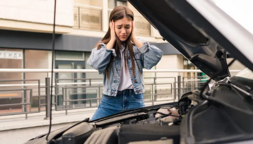 Zrozpaczona dziewczyna z rękami na uszach przy otwartej masce samochodu. Jej auto nie odpala, co ją bardzo frustruje.