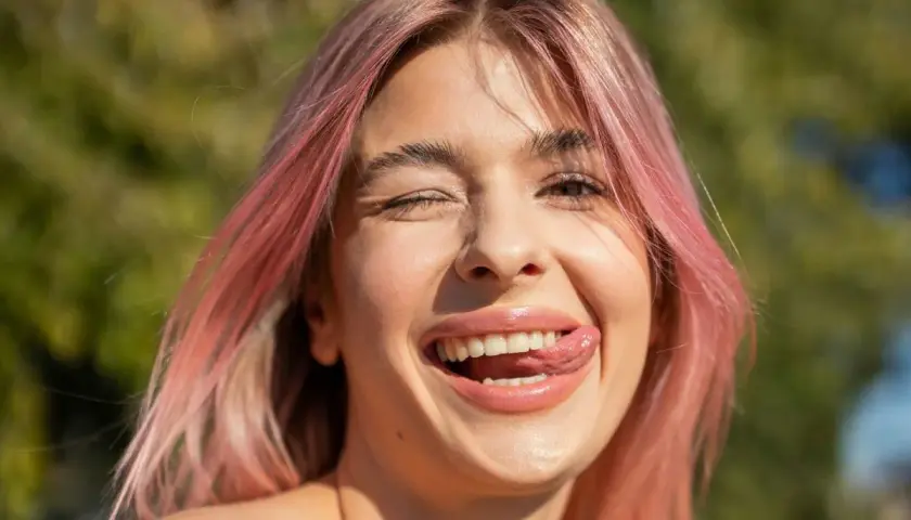 Uśmiechnięta dziewczyna z truskawkowym blond włosem puszcza oczko i wystawia język.