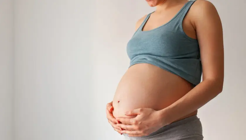 Uśmiechnięta ciężarna kobieta trzyma dłonie na brzuchu. Czas na poród zbliża się, może już niedługo pojawi się czop śluzowy.
