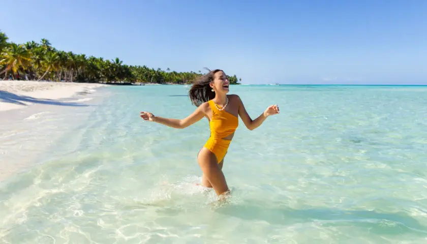 Szczęśliwa kobieta w żółtym stroju kąpielowym biegnie przez turkusową wodę na plaży z palmami w tle.