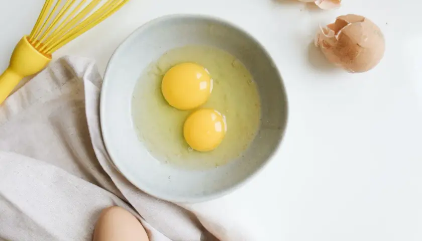 Deux jaunes d'œufs dans un bol, prêts à être battus. Une question se pose : peut-on congeler des jaunes d'œufs ? Un fouet jaune et des coquilles d'œufs complètent la scène.