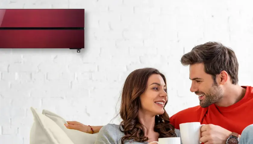Pareja sonriente en un sofá, disfrutando de café. Arriba, un aire acondicionado rojo moderno garantiza cop eficiencia.