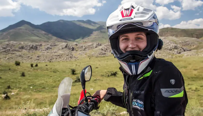 Uśmiechnięta motocyklistka w kasku, gotowa do podróży. W tle górzysty krajobraz, idealny do przygód z **carnet de passage**.