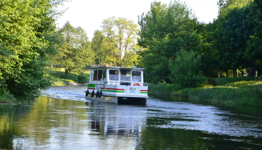 Biały tramwaj wodny Gordalina płynie po spokojnej rzece, otoczony zielenią drzew i traw.
