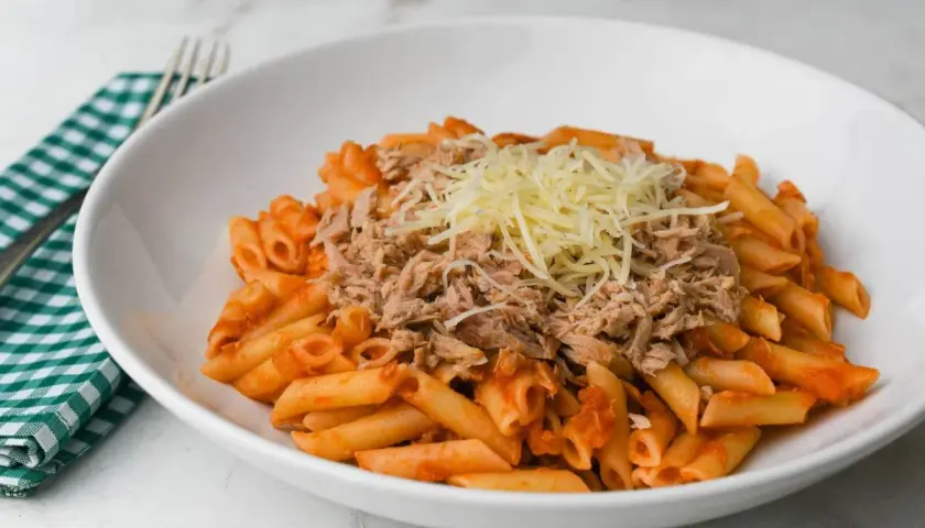 Macarrones con tomate y atún desmenuzado, cubiertos con queso rallado, servidos en un plato blanco.