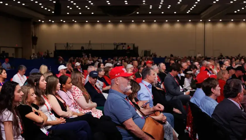 A large audience attends a government conference, with many people wearing red hats.