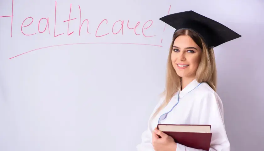Laurea in medicina ma non voglio fare il medico. Studentessa con tocco e camice, libro in mano, davanti a una lavagna con la scritta "Healthcare".