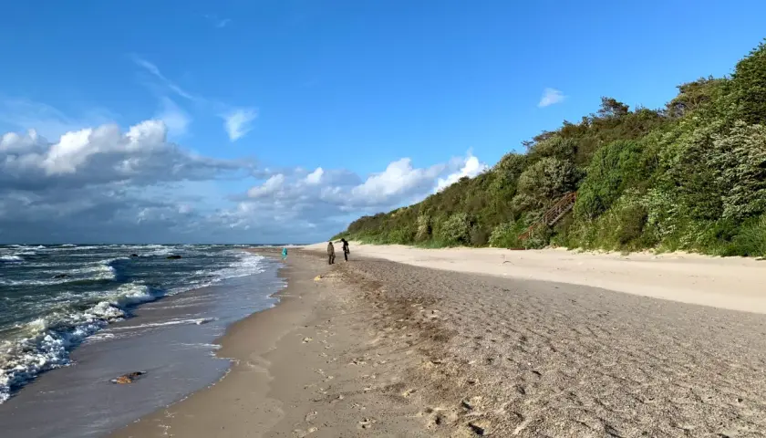 Spaziergang am Strand von Dranske Bakenberg. Wellen brechen an der Küste, während zwei Personen den Sandweg entlanggehen.