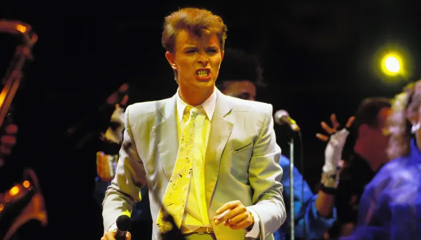 David Bowie na scenie podczas Live Aid, w jasnej marynarce i żółtym krawacie, śpiewa z pasją.