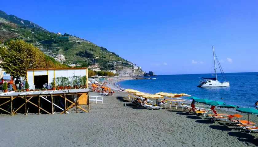 Plage de sable noir à Maiori, Italie, avec parasols, chaises longues, un catamaran blanc et des collines verdoyantes.