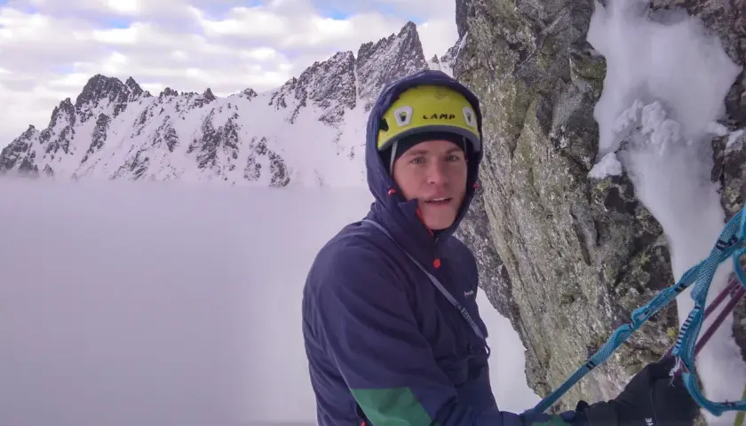 Alpinista con casco amarillo y ropa de montaña, rodeado de picos nevados y niebla.