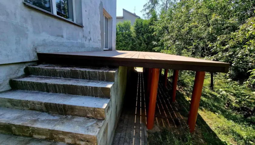Nowy, wysoki taras na słupach, z betonowymi schodami prowadzącymi do niego. Otoczony zielenią.