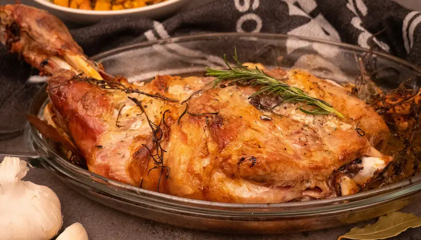 Une succulente épaule d'agneau rôtie, garnie de romarin frais, dans un plat en verre. A côté, de l'ail et une purée de légumes.