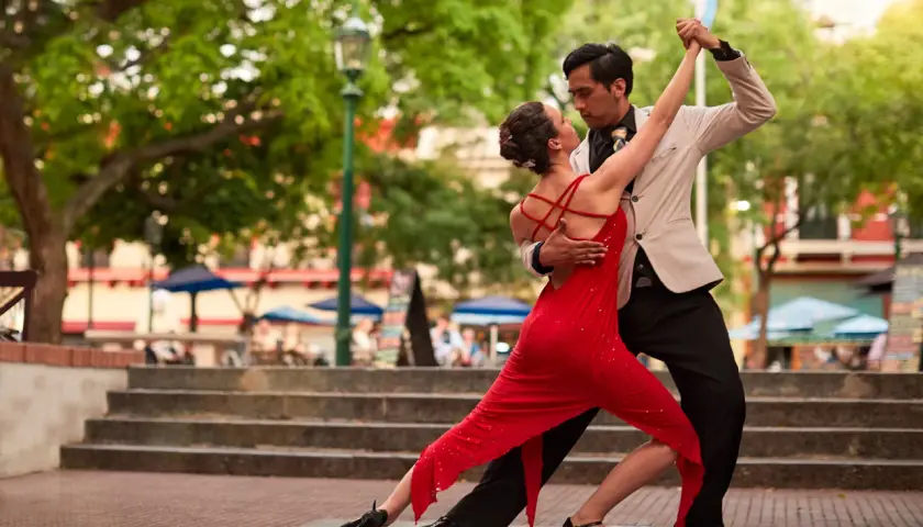 Coppia di ballerini latino americani in un appassionato tango all'aperto, lei in abito rosso, lui in giacca chiara.