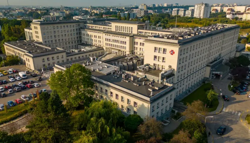 Widok z lotu ptaka na duży kompleks szpitalny z czerwonym krzyżem na fasadzie, otoczony drzewami i parkingiem.