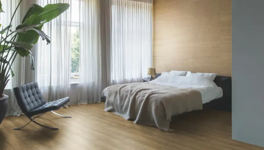 Sypialnia z łóżkiem, fotelem i rośliną. Podłoga z paneli ułożona równolegle do okna, co optycznie powiększa przestrzeń.