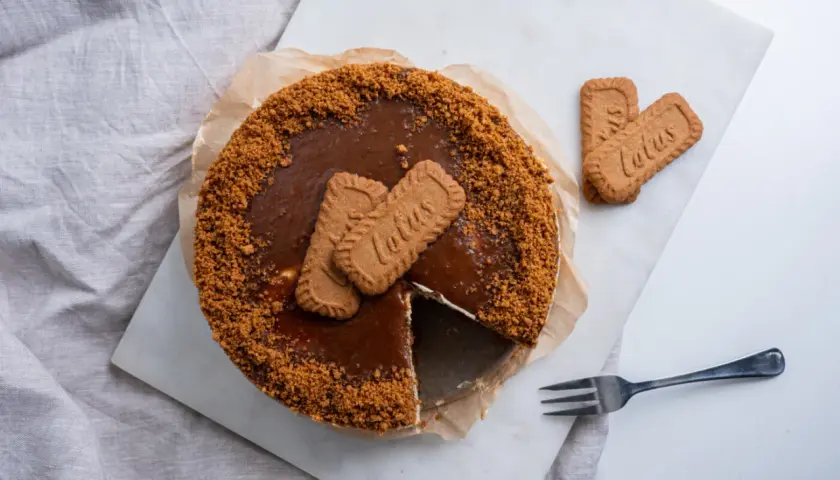 Pyszny sernik lotus z polewą karmelową i pokruszonymi ciasteczkami. Obok leżą dwa ciasteczka lotus i widelec.