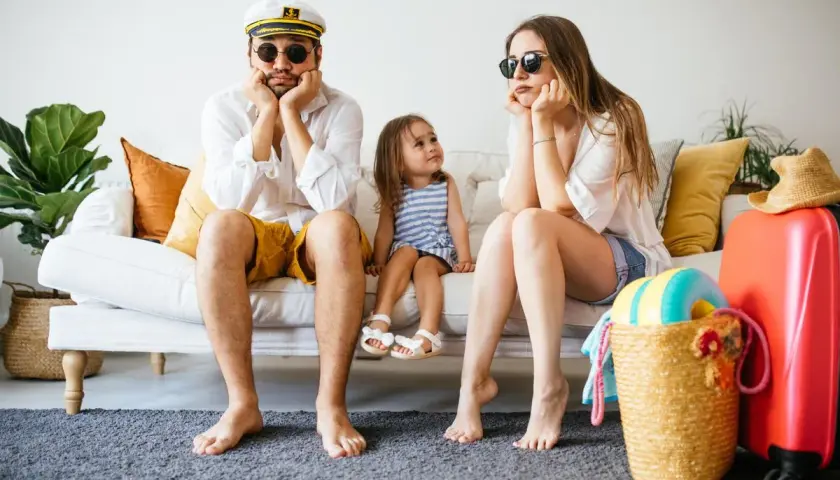 Famille dépitée sur un canapé, valise rouge prête. Annulation location vacances cas de force majeure, les vacances rêvées s'envolent.