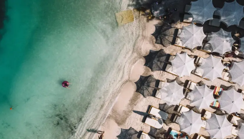 Widok z lotu ptaka na turkusowe morze i piaszczyste plaże Albanii. Jedna osoba relaksuje się na dmuchanym kole, a obok rzędy białych parasoli i leżaków.