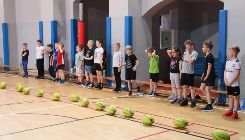Dzieci w wieku 4-6 lat stoją w rzędzie na sali gimnastycznej, gotowe do gier i zabaw ruchowych. Na podłodze leżą piłki.