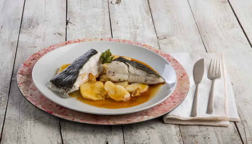 Rodaballo gallego servido con patatas y salsa, acompañado de cubiertos y servilleta sobre una mesa de madera.