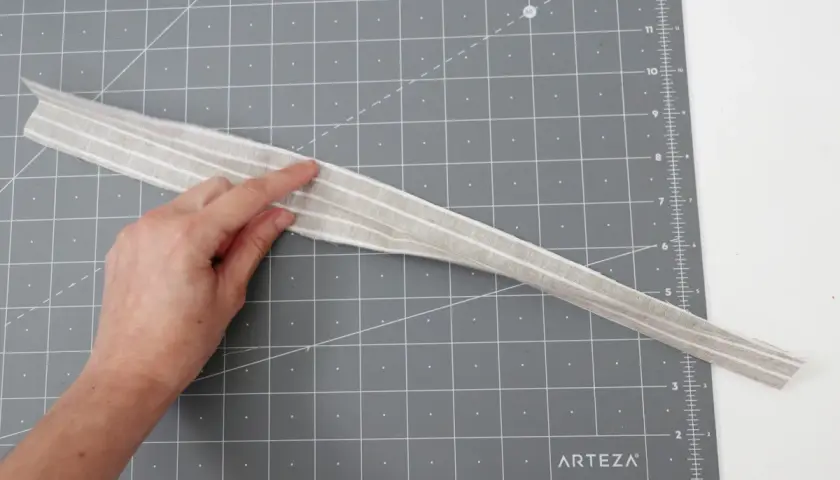 A hand points to a striped fabric strip, possibly for a belt loop pattern, on a cutting mat.