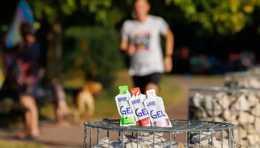 Trzy opakowania żeli energetycznych dla biegaczy: zielone jabłko, cola i truskawka z bananem, na tle biegnącej osoby.
