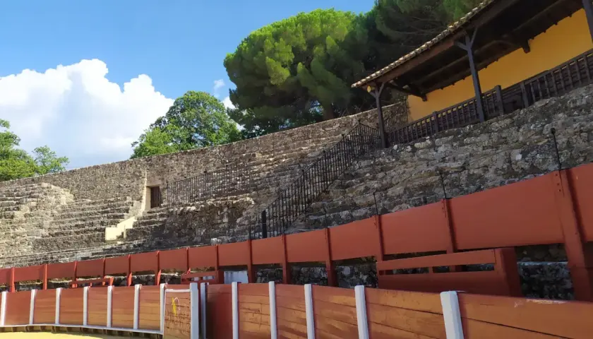 La plaza de toros cuadrada en España, con sus gradas de piedra y barreras de madera, espera la emoción bajo un cielo azul y nubes algodonosas.