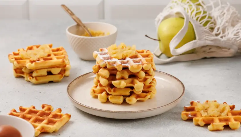 Goldbraune Waffeln mit Apfelmus bestreut, daneben eine Schüssel mit Apfelmus und ein grüner Apfel im Netzbeutel.
