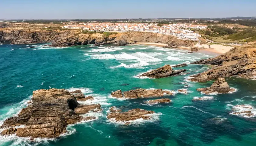 Costa rocosa y aguas turquesas en una **playa Portugal**. Un pueblo blanco se asoma al acantilado.