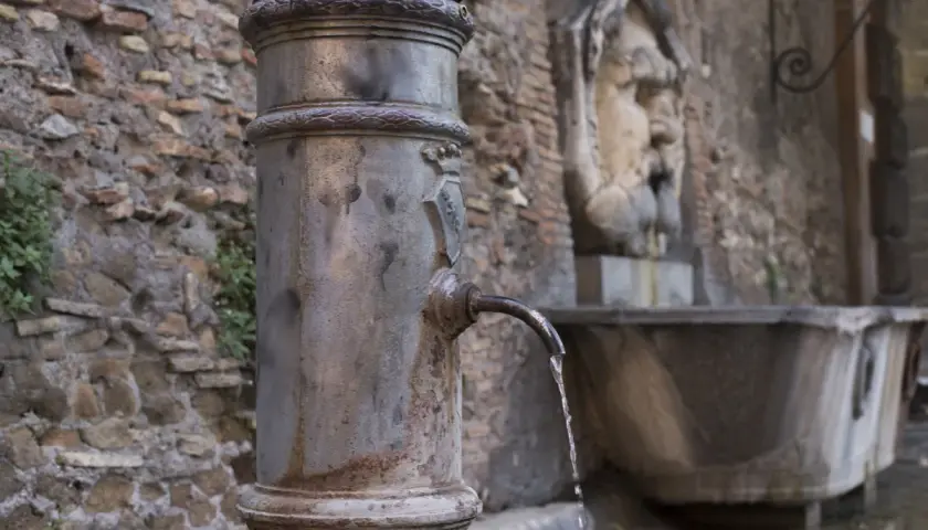 Fontanella romana con un nasone che eroga acqua fresca. Sullo sfondo, un antico volto scolpito.
