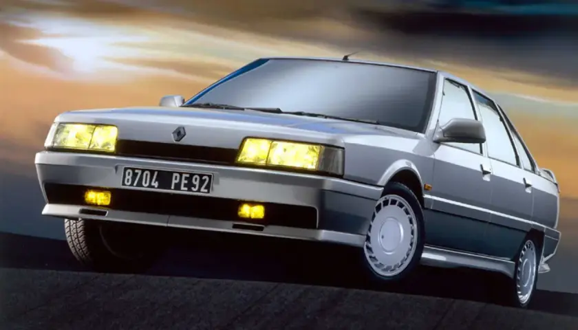 Un elegante Renault 21 Turbo plateado, con sus faros amarillos encendidos, se luce contra un cielo crepuscular.