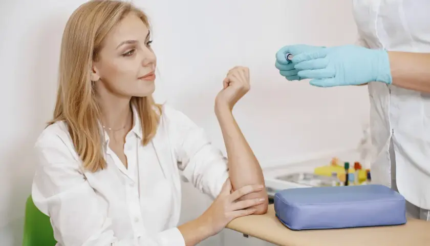 Kobieta z blond włosami w białej koszuli i pielęgniarka w rękawiczkach przygotowują się do pobrania krwi.