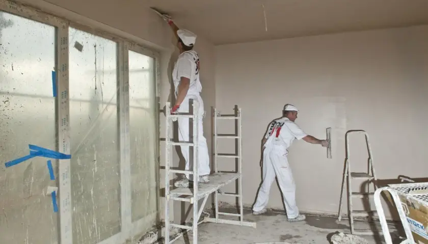 Dwóch fachowców w białych kombinezonach, używając kielni, wyrównuje ściany gładzią.