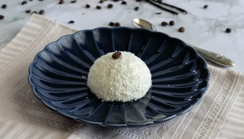 Semifreddo tartufo bianco, un dolce raffinato ricoperto di cocco e decorato con un chicco di caffè, su un piatto blu scuro.