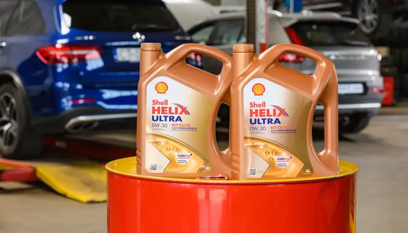 Dwa kanistry oleju silnikowego Shell Helix Ultra 0W-30 stoją na czerwonym bębnie. W tle widać samochody w warsztacie.