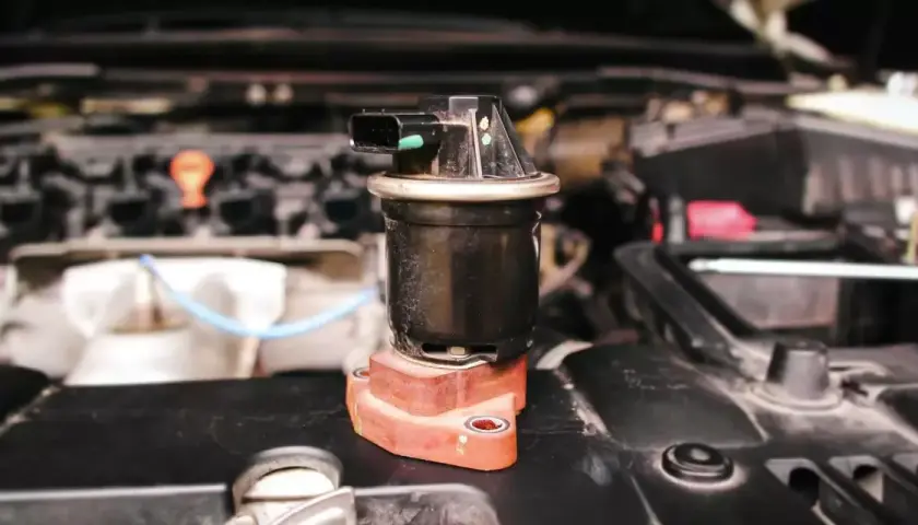 Componente negro con conector eléctrico, posiblemente una válvula de admisión, sobre una base rojiza en un motor.